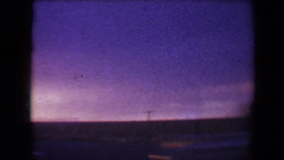 1967 8mm frame from Flagstaff, Arizona, with a purple evening sky, dark horizon, and faint passing-car light streaks near the bottom edge.