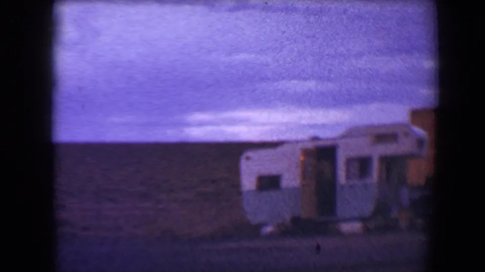 The 1967 Flagstaff home-movie frame shows a small blue and white camper parked on a roadside with low hills behind it.