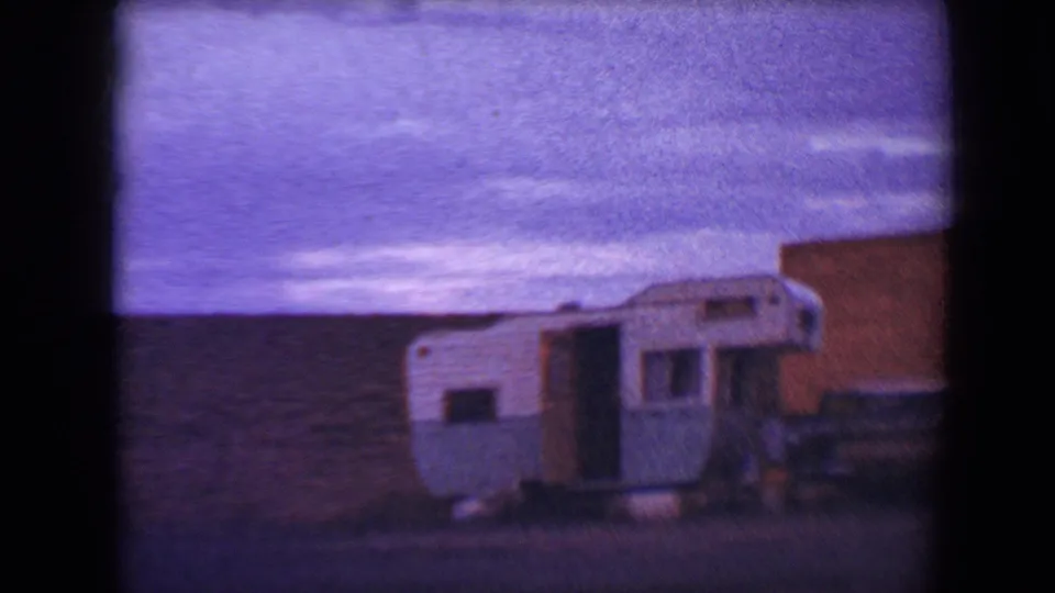 A blue and white camper is framed beside a remote Flagstaff road, with open ground, low hills, and a purple color cast from the 8mm film.