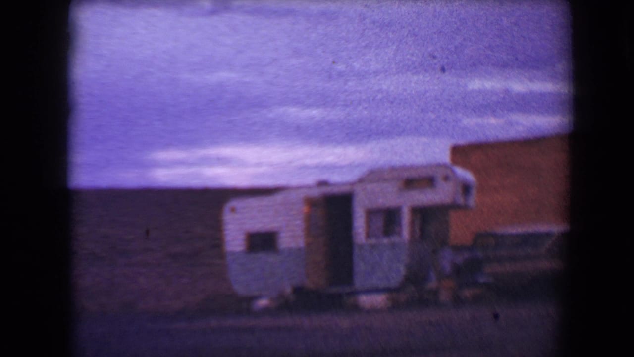 1967 Flagstaff home-movie frame showing a white and blue travel trailer with its door open near a brick drive-in structure.