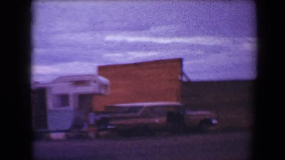 1967 Flagstaff home-movie frame showing the trailer partly at left, a brick drive-in structure, and a dark station wagon near the curb.