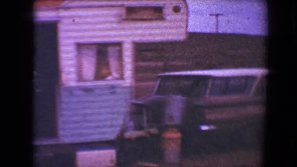 1967 Flagstaff home-movie frame with the trailer window, striped siding, and a parked station wagon in soft purple-tinted film color.