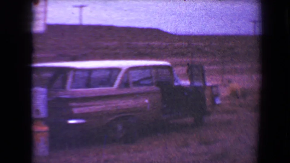 1967 8mm frame from Flagstaff showing a vintage station wagon beside a travel trailer against low rural hills.