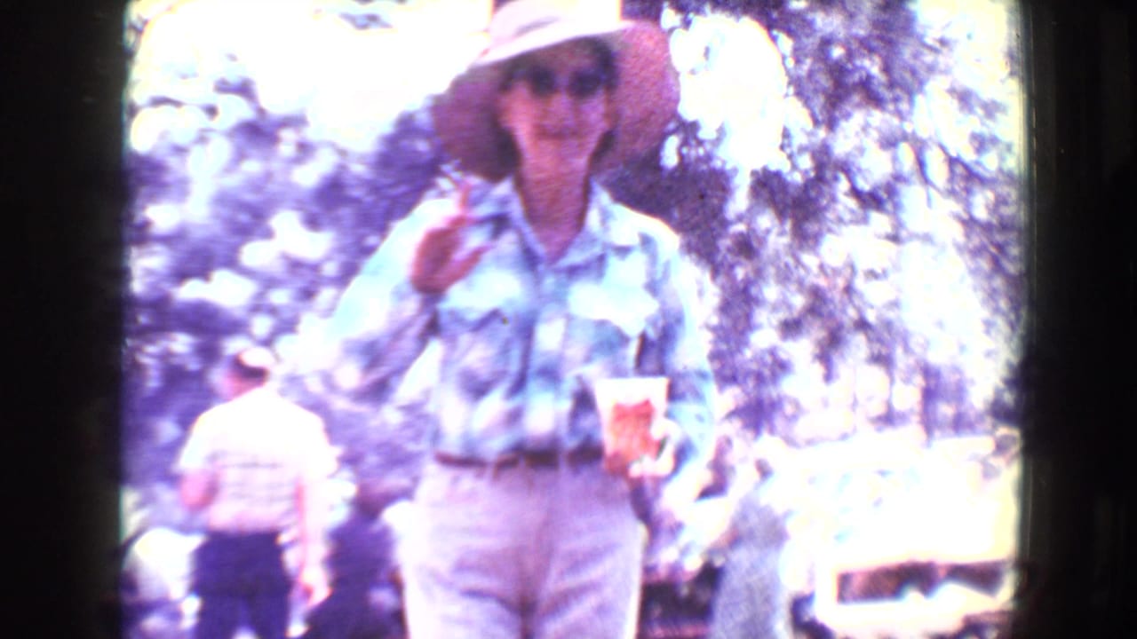 In 1967 Flagstaff home-movie footage, a woman in casual clothing walks toward the camera holding a drink near seated adults and pine trees.