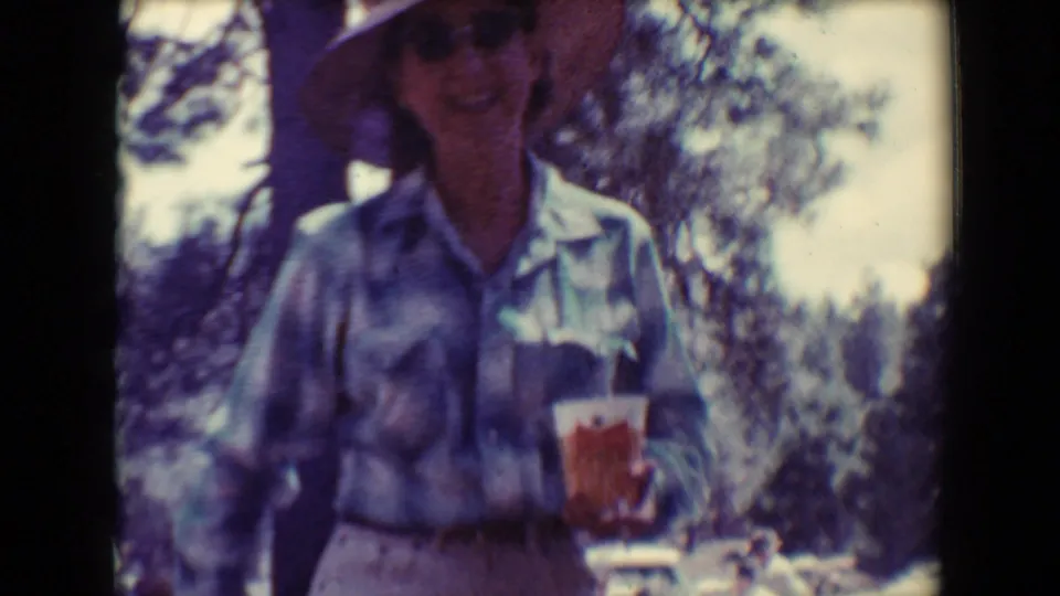 A woman in a wide-brimmed hat and plaid shirt holds a red drink container in a wooded Flagstaff, Arizona home-movie scene.