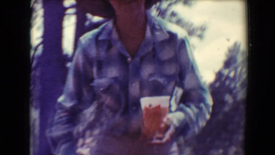 Closer 8mm frame of the woman in a plaid shirt holding a drink, with trees and another standing figure partly visible behind her.