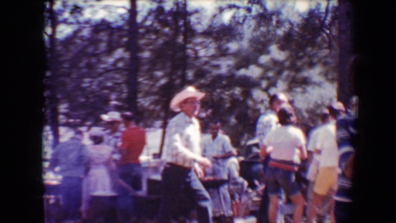 Adults gather near barbecue tables in a wooded outdoor area with smoke at left.
