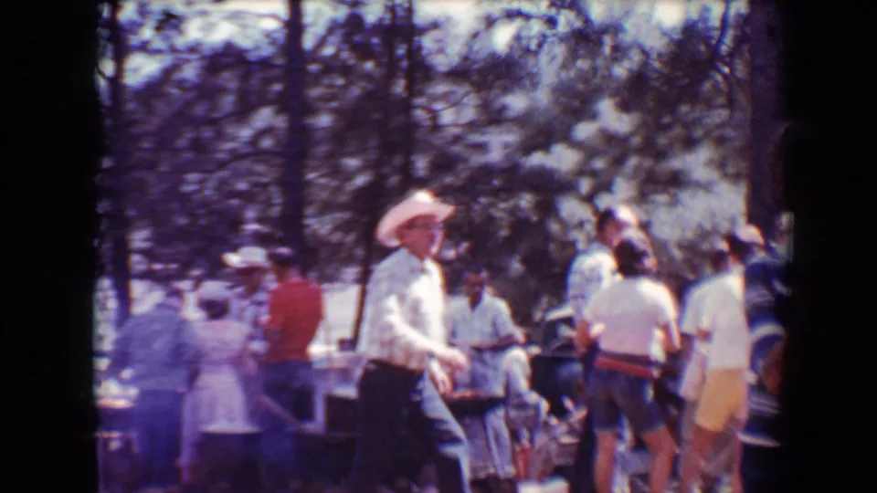 1967 Flagstaff home-movie frame showing adults in summer clothing near barbecue tables among trees.