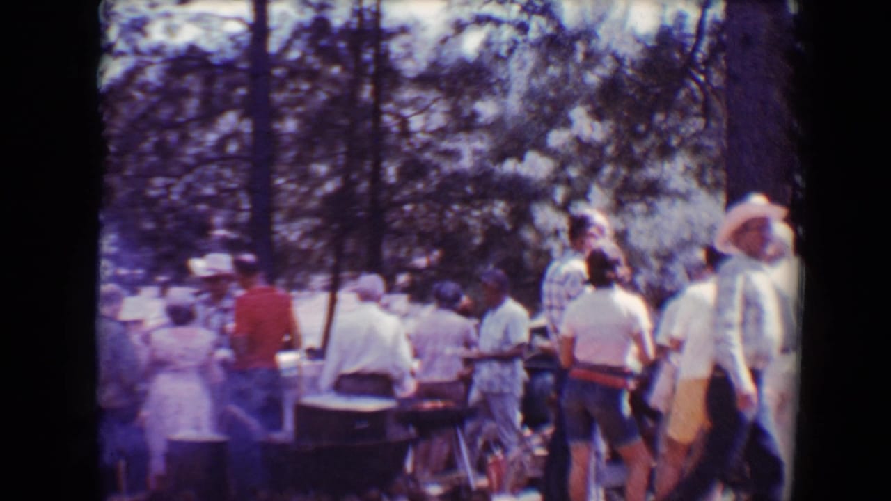 Adults in casual 1960s clothing stand around barbecue tables in a wooded Flagstaff outdoor setting.