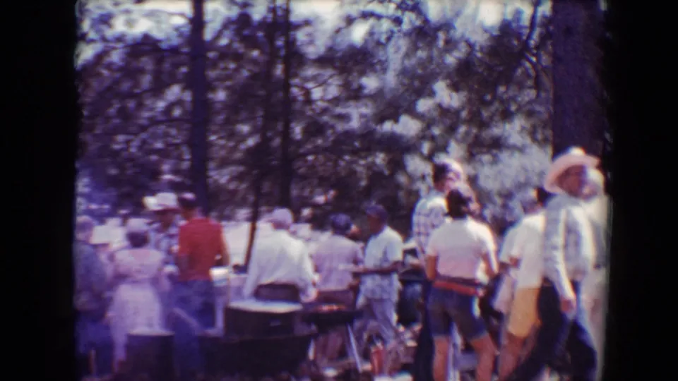 Adults in casual 1960s clothing stand around barbecue tables in a wooded Flagstaff outdoor setting.