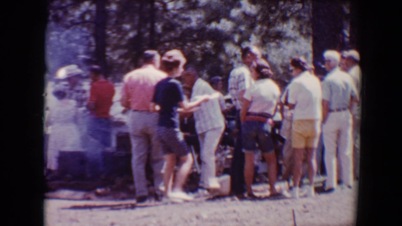 Adults stand in line near a smoky outdoor barbecue in a wooded park.