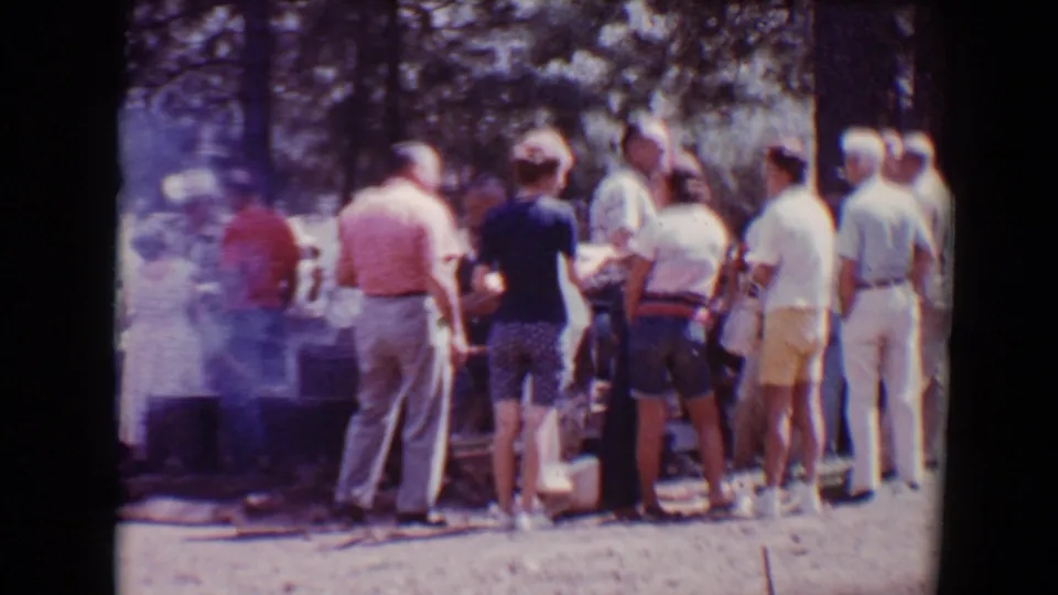 Adults gather in a loose food line beside barbecue smoke in 1967 Flagstaff home-movie footage.