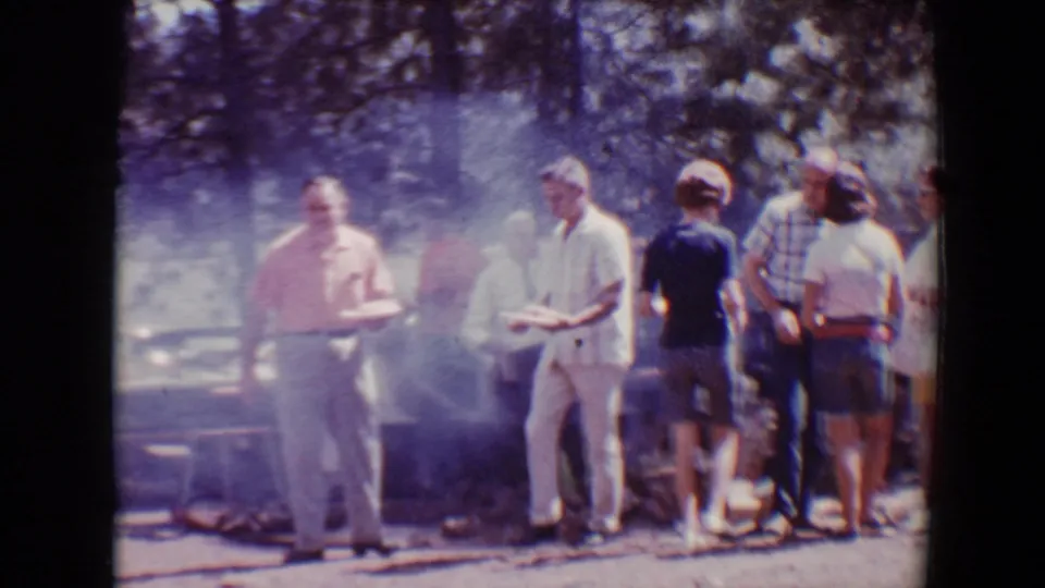 Adults holding plates stand near drifting barbecue smoke in a 1967 Flagstaff outdoor gathering.