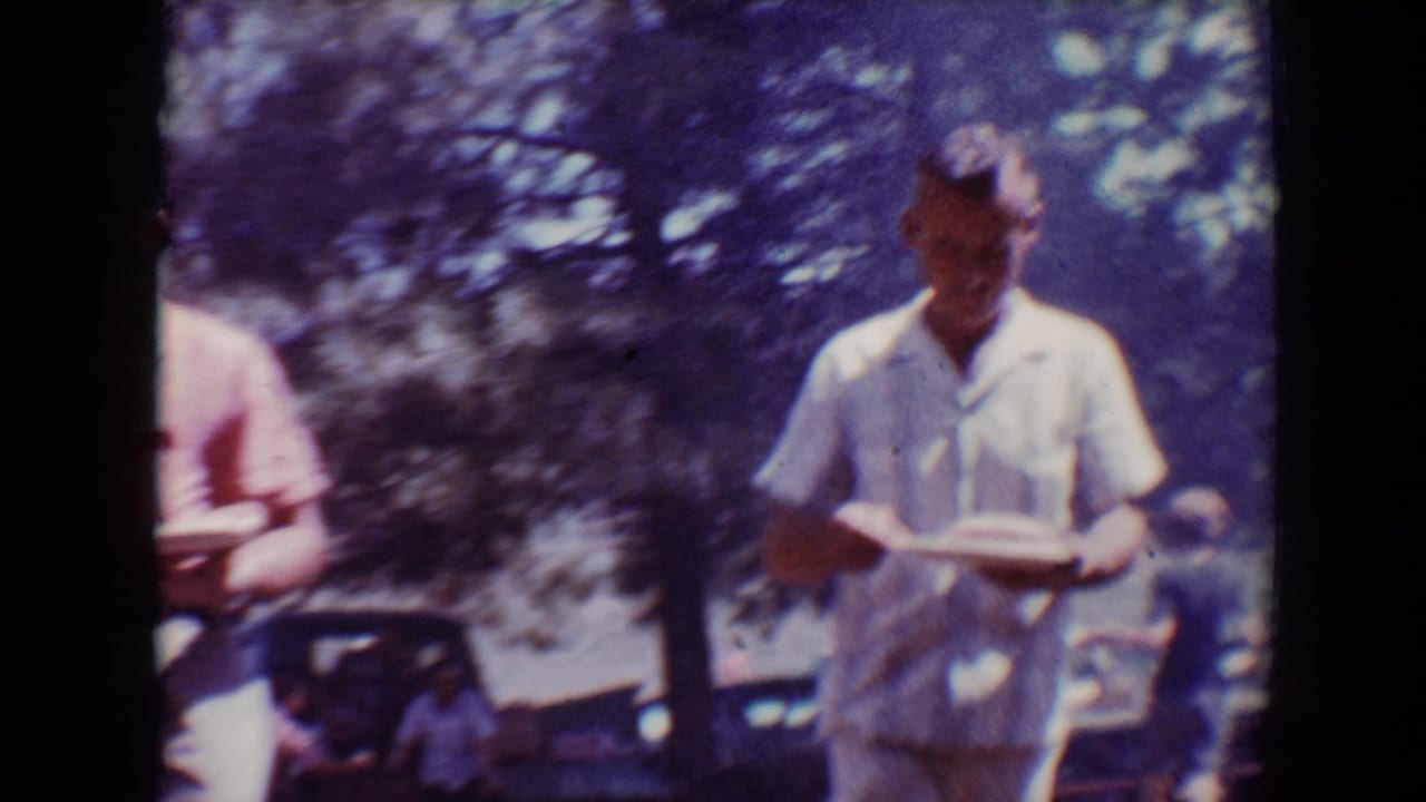 In 1967 Flagstaff home-movie footage, a man in a white short-sleeve shirt walks toward the camera holding a plate in a wooded outdoor area.