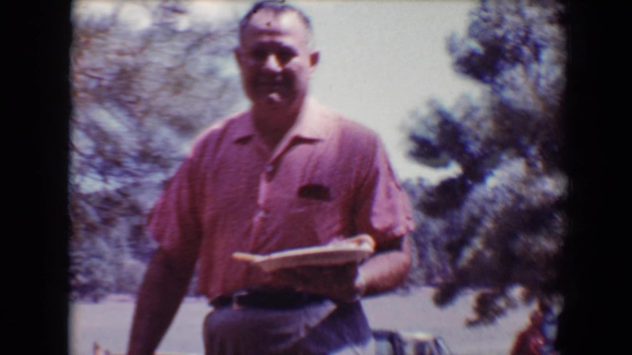 Man in a pink shirt smiles while walking outdoors with a plate in his hand.