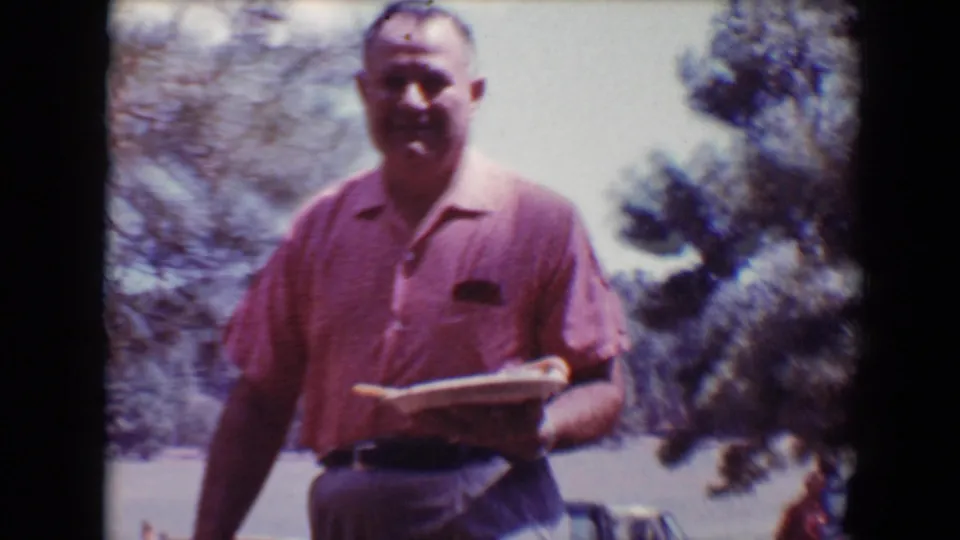 A man in a pink short-sleeve shirt smiles toward the camera while holding a plate in a sunny wooded Flagstaff setting.