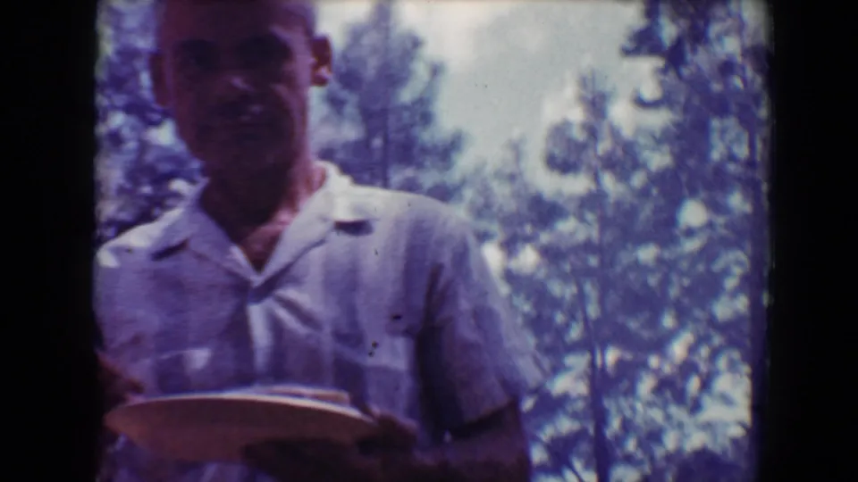 Close home-movie view of an adult man in a light patterned shirt holding a plate, with sunlit trees behind him in Flagstaff.
