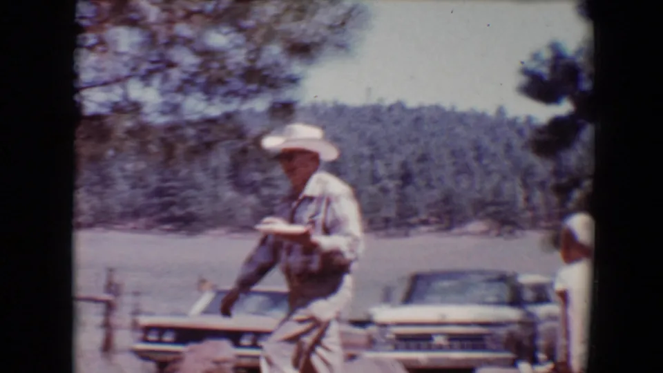 A man wearing a cowboy hat and jacket carries a plate near vintage parked cars, with wooded hills visible in the Flagstaff background.