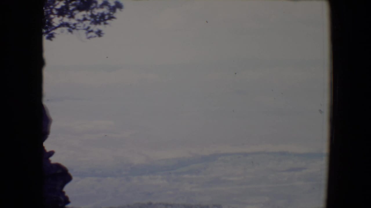 1967 8mm home-movie view near Flagstaff, Arizona, with dark rocks and tree branches framing distant hazy ridges.