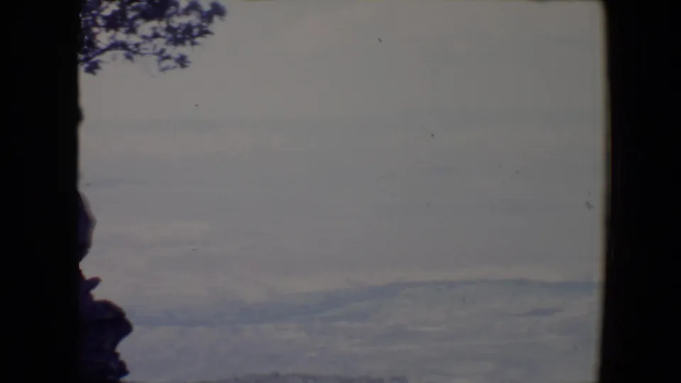 1967 8mm home-movie view near Flagstaff, Arizona, with dark rocks and tree branches framing distant hazy ridges.