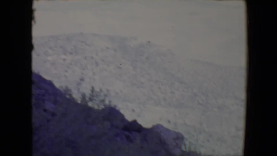 1967 8mm Flagstaff landscape view with dark foreground rocks, a forested slope, and a pale overcast sky.