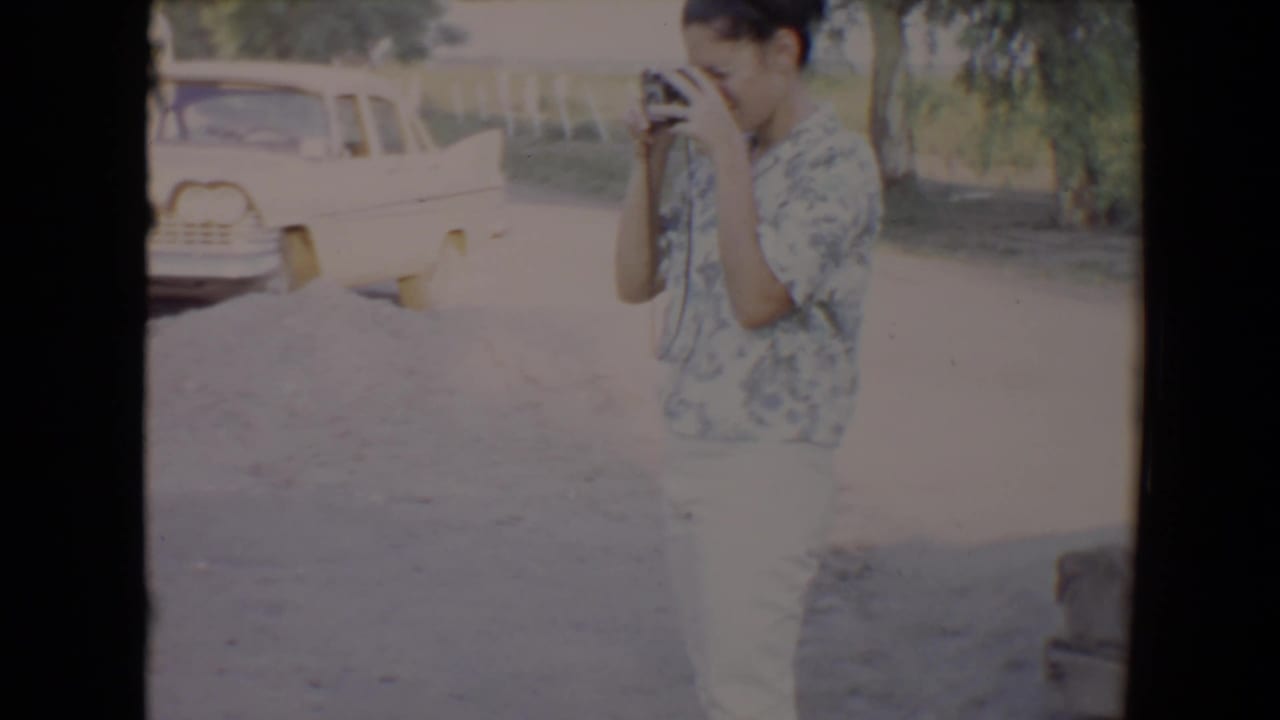 In 1967 Flagstaff home-movie footage, a woman in a patterned shirt appears to take a photo beside a parked light-colored car.
