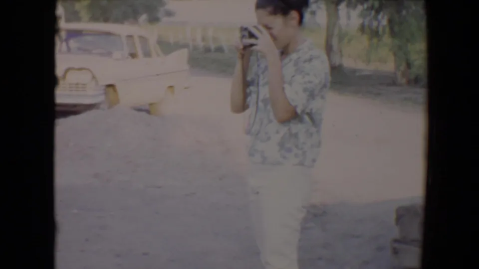 In 1967 Flagstaff home-movie footage, a woman in a patterned shirt appears to take a photo beside a parked light-colored car.