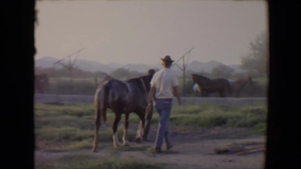 1967 Flagstaff frame showing a man in a white shirt and cowboy hat leading a horse across a rural field with other horses and mountains beyond.