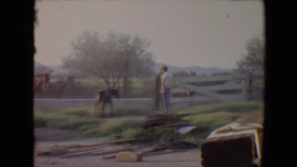 1967 Flagstaff frame with a man in a hat beside a white fence as horses stand in a grassy rural area.