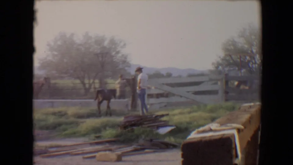 1967 Flagstaff home-movie frame showing a man near a pony and another figure beside a white fence in a rural paddock.