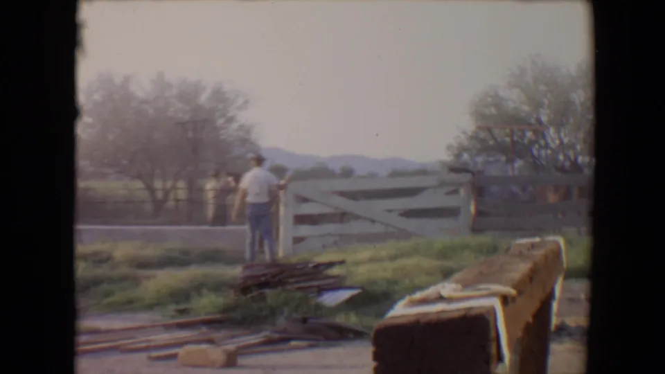 1967 Flagstaff frame showing a man in a cowboy hat and another person near a white wooden gate with rural hills behind them.