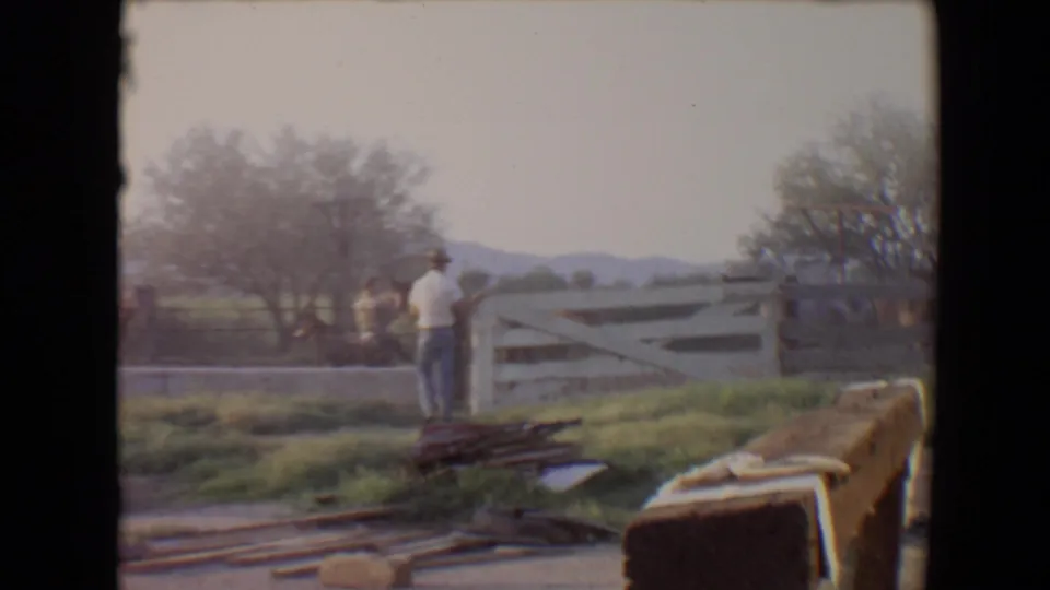 1967 Flagstaff home-movie frame with a man by a white fence watching horses in a grassy paddock with trees and mountains beyond.