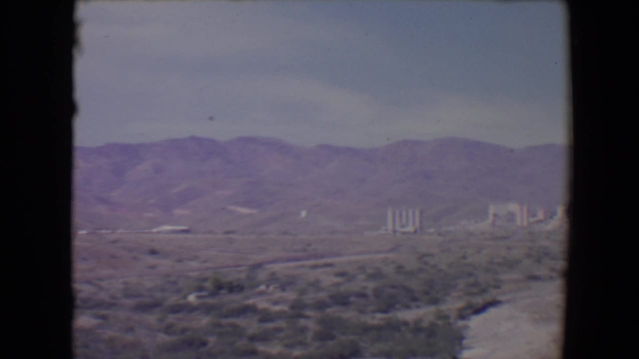 Desert landscape with mountains, pale sky, scrub vegetation, and distant industrial buildings.