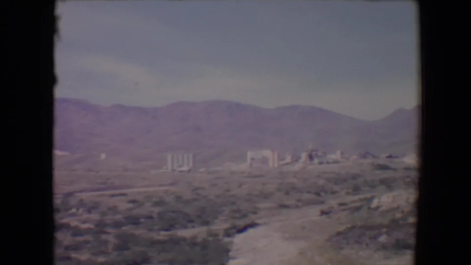 Faded 8mm frame from 1967 near Flagstaff showing a broad arid foreground, mountains, and distant industrial buildings.