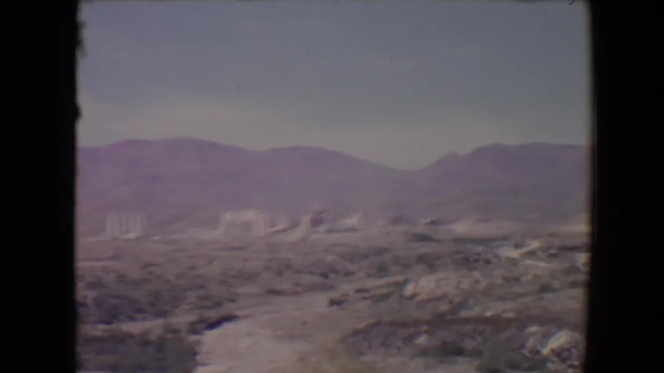 Soft 1967 8mm view near Flagstaff with arid plants and rocks in front of mountains and industrial buildings.