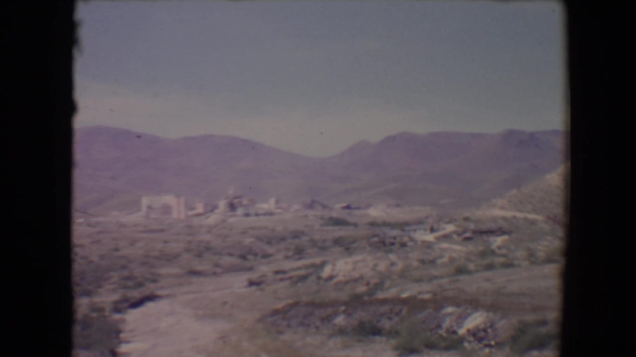 1967 8mm view near Flagstaff showing arid hills, distant low buildings, mountains, and a pale sky.