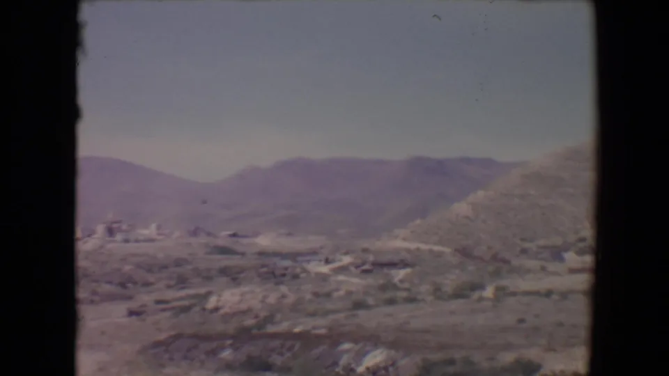 Flagstaff-area 1967 home-movie frame with dry terrain, distant settlement, mountains, and a dark speck in the sky.