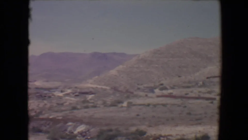 1967 8mm frame near Flagstaff showing a rounded dry hill, scrub vegetation, and distant mountain slopes.