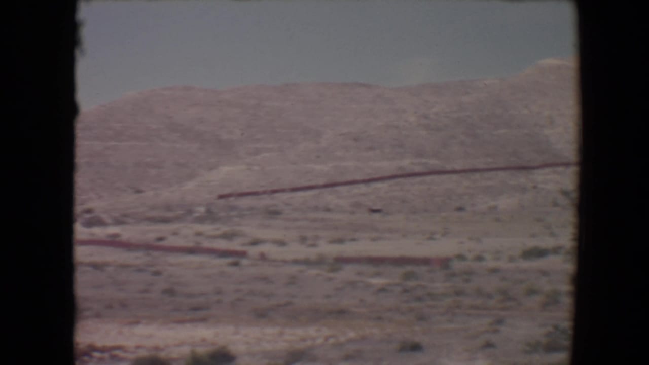 Desert hillside with sparse scrub and a red linear structure under a hazy sky.
