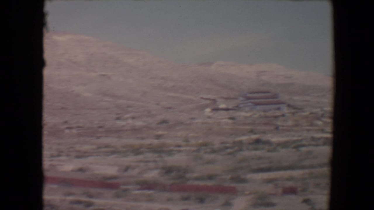 Pale desert mountainside with scrub and distant low red-roofed structures under a hazy sky.