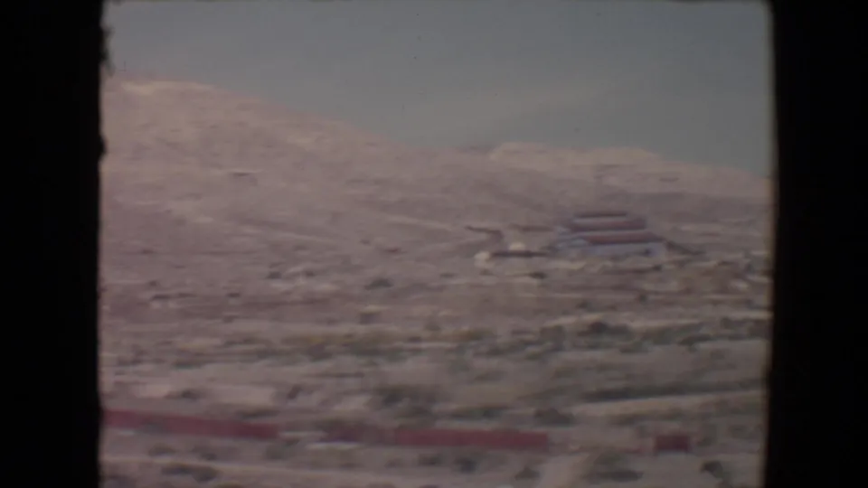 1967 8mm view near Flagstaff, Arizona, showing a pale scrub-covered slope and distant red-roofed structures from a moving vehicle.