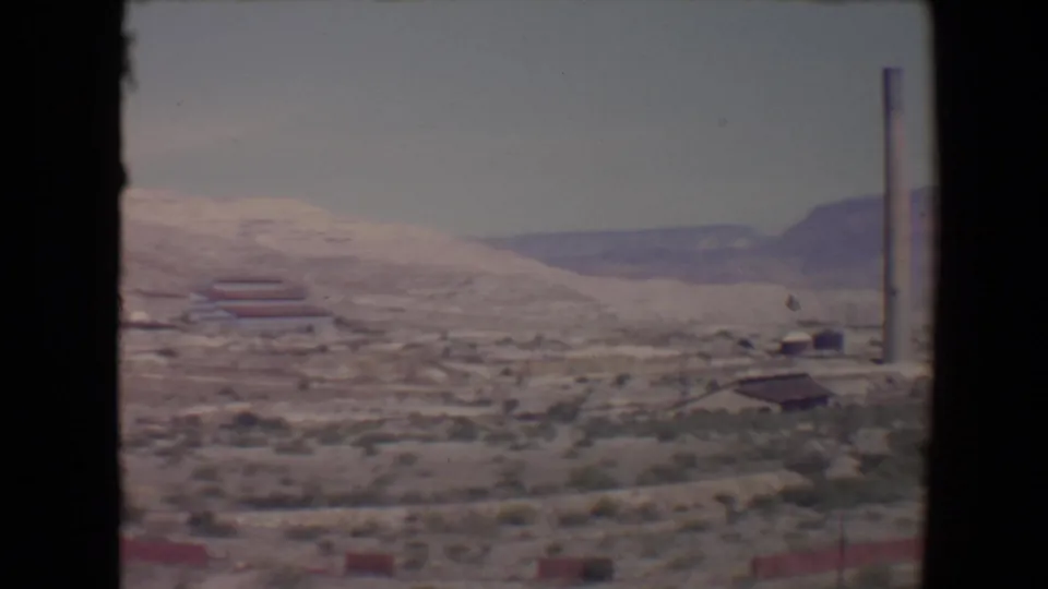 1967 Flagstaff-area 8mm frame with scrubby terrain, distant low structures, a tall smokestack, and purple-brown ridgelines.