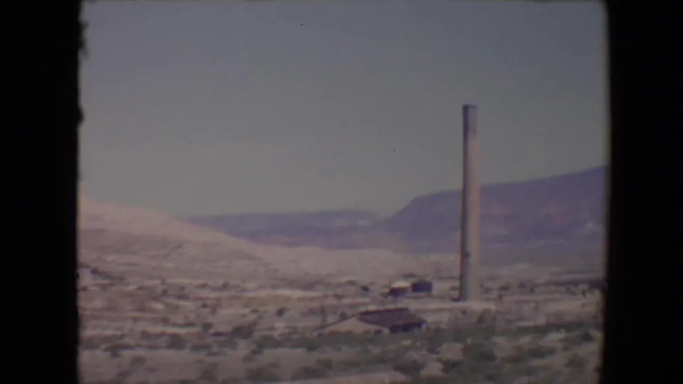 1967 8mm roadside view near Flagstaff with a smokestack, low red-roofed structure, pale hillside, and distant mesa-like ridges.