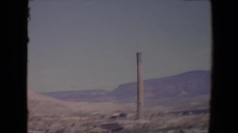 Soft 1967 8mm frame near Flagstaff showing a tall smokestack, small distant building, scrubby foreground, and muted purple ridges.