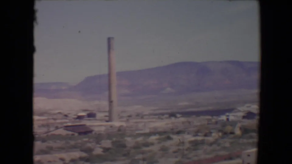 Flagstaff, Arizona home-movie frame showing a smokestack, scattered structures, scrubby ground, and purple-toned hills.