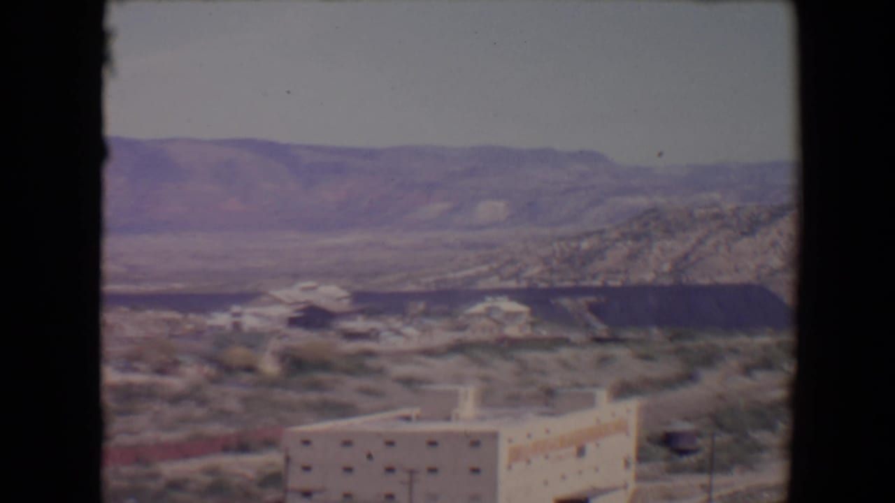 Desert valley with a pale multi-storied building, scrub brush, roads, distant hills, and a dark excavation.