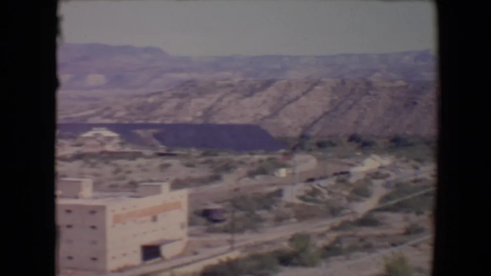 Flagstaff, Arizona home-movie frame showing a valley building, scattered development, and a dark excavation below rugged hills.