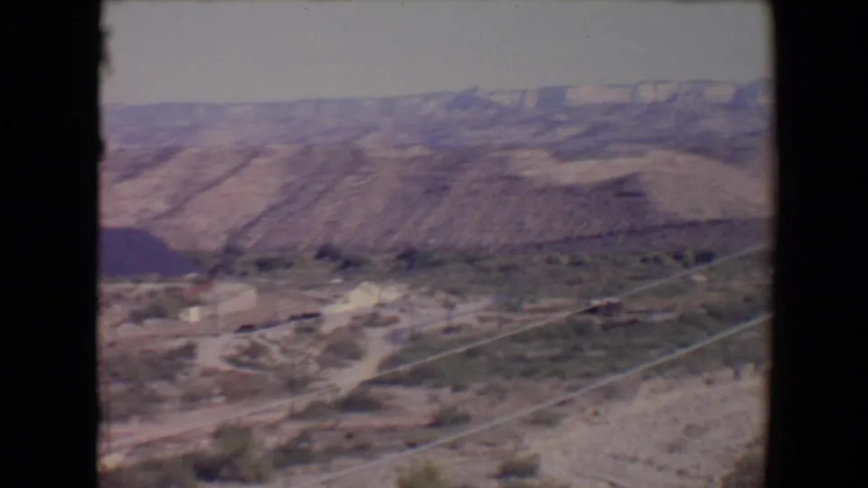 1967 8mm view across a Flagstaff-area valley with sparse development, pale tracks, brush, and layered hills.