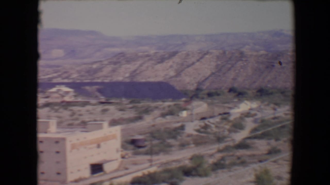 1967 8mm view over Flagstaff, Arizona, showing pale buildings, dry terrain, and layered mountains in the distance.