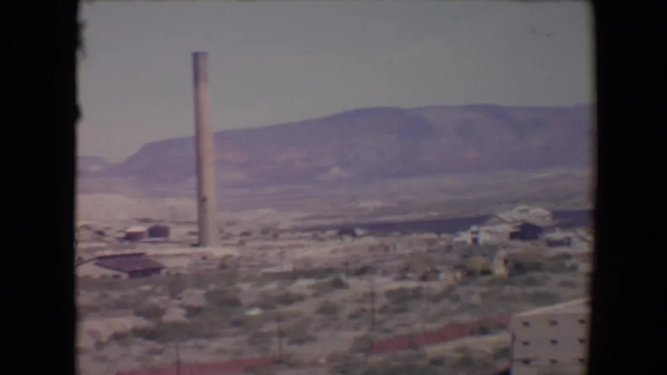 1967 Flagstaff view with a tall smokestack, low industrial buildings, scattered trees, and mountains beyond.